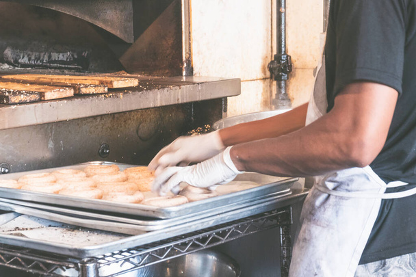 США, Нью-Йорк: Традиционная выпечка багеля. Пекарь в процессе приготовления подлинных бубликов нью-йоркского стиля с кунжутом для выпечки в духовке
. - Фото, изображение