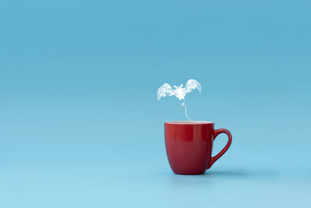 Steam in bat shape flying from coffee cup against blue background. Morning drink. Halloween celebration concept. Copy space. - Φωτογραφία, εικόνα