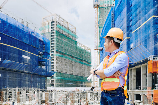 Souriant travailleur de la construction asiatique avec le plan dans sa main en tournant le dos et en regardant le bâtiment en construction
 - Photo, image