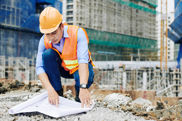 Serious Vietnamese contractor in orange hardhat examining building blueprint when working at construction site - Foto, Imagem