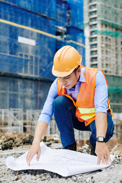 Профессиональный азиатский инженер-строитель, распространяющий план строительства на земле и изучающий план этажа
 - Фото, изображение