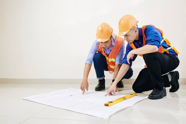 Engineers in hard hats and orange vests discussing floor layout in big blueprint on the floor - Photo, Image