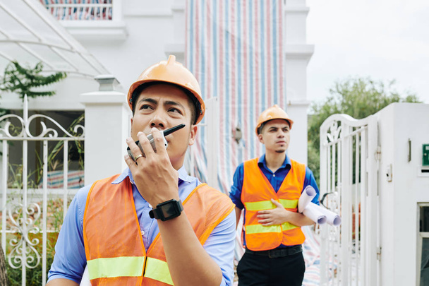 Serious frowning Vietnamese construction engineer talking on walkie-talkie when he and his coworker leaving building they were working in - Photo, Image