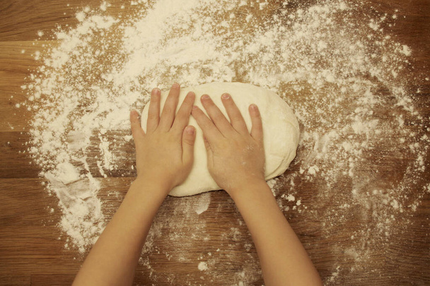 children's hands knead the dough on a wooden table, sprinkled with flour, close-up, selective focus, home cooking concept, copy space - Photo, Image