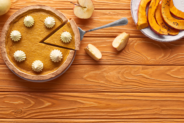 delicious pumpkin pie with whipped cream near cut apple, and sliced baked pumpkin on orange wooden table - Foto, afbeelding