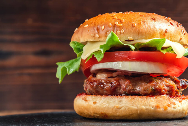 Fresh homemade burger with meat cutlet, tomatoes, lettuce, cheese and onions on a slate dish on a wooden background - Foto, Imagen