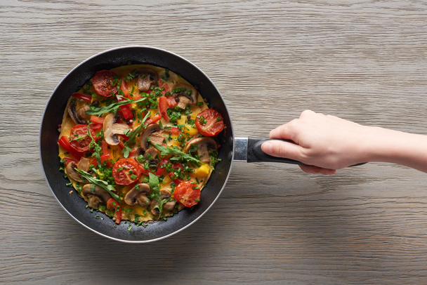 cropped view of woman holding frying pan with omelet with mushrooms, tomatoes and greens - Valokuva, kuva
