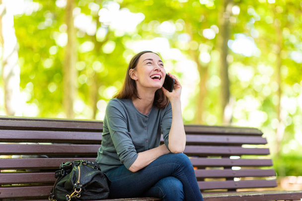Portrait happy woman sitting on park bench talking with cellphone - Foto, Imagem