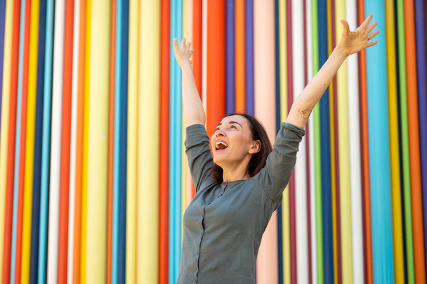 Portrait  cheerful middle aged woman with arms raised by colorful background - Photo, Image