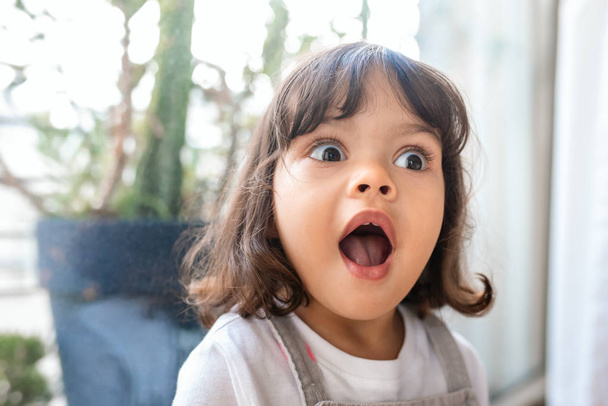 Adorable little girl with her mouth and eyes wide open in surprise while playing in her living room at home - Foto, afbeelding