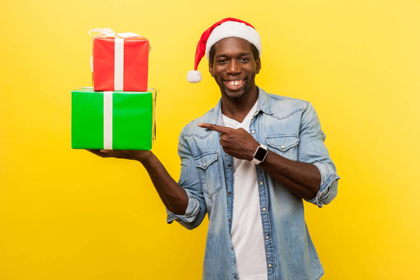 positive handsome young man in santa hat and casual denim shirt pointing at gift boxes on hand and happy looking at camera  on yellow background, xmas shopping concept  - Foto, immagini