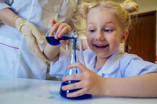 surprised and amazed clever blonde school girl chemist in eyeglasses and white medical gown making science experiments chemistry ,mixing different chemical solutions in laboratory classroom - Fotografie, Obrázek