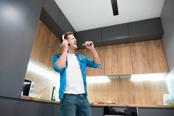 νεαρός όμορφος άντρας με ακουστικά χορεύει ενώ μαγειρεύει στην κουζίνα - Φωτογραφία, εικόνα
