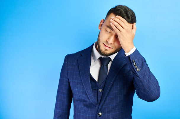 Asian Kazakh man in business suit holds his head, experiencing stress and headache at office work, tired of fuss isolated on blue studio background - Valokuva, kuva