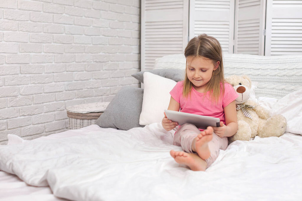 cute little girl lies in bed uses digital tablet. - Фото, изображение