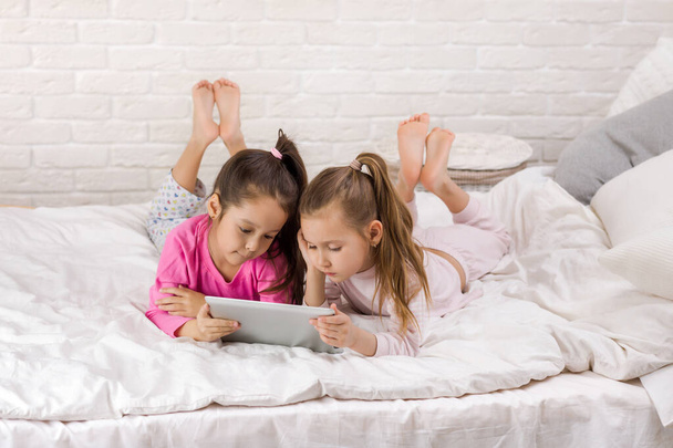 two cute children girls use digital tablet in bed. - Фото, изображение
