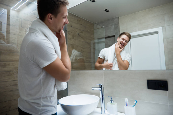 young handsome smiling man with white towel on his shoulders looking in the bathroom mirror - Фото, изображение
