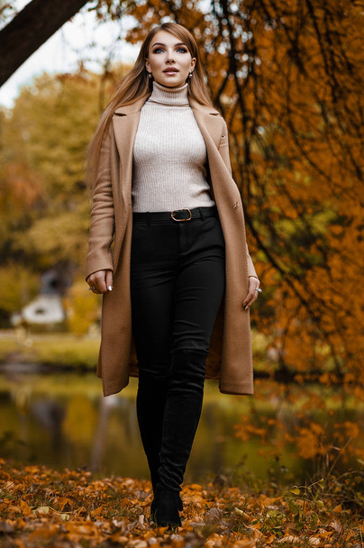 A blonde in an autumn beige coat is walking in the park. Seasonal photo. Glamor Fashion - 写真・画像