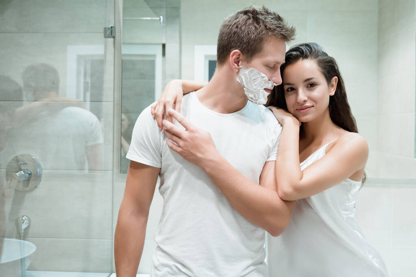 young couple handsome man and beautiful woman hugging in bathroom while he has shaving foam on his face - 写真・画像