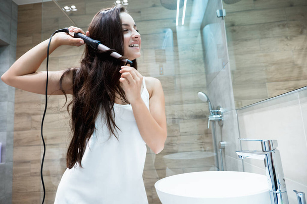 young beautiful smiling woman in white gown standing in the bathroom curling her long brunette hair with iron - Fotó, kép