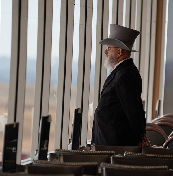 Older man in turn of the century suit and top hat, stands in front of the windows overlooking the race track at Arizona Downs. - Photo, Image