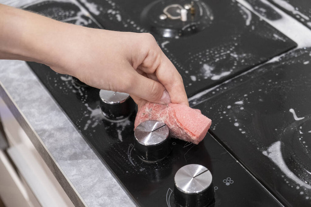 Female hand washes gas stove, glass ceramic surface. Red sponge and white foam. - 写真・画像