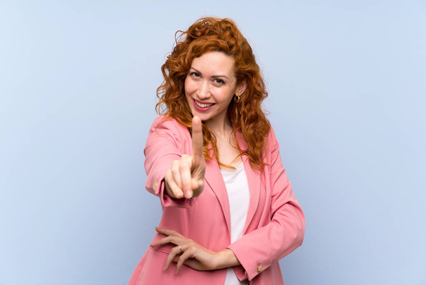 Mulher ruiva de terno sobre parede azul isolada mostrando e levantando um dedo
 - Foto, Imagem