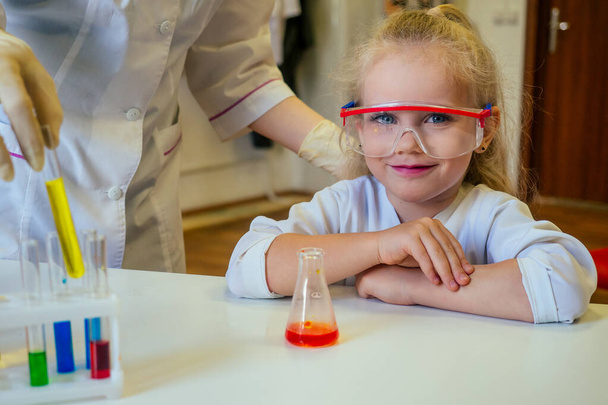clever blonde school girl chemist in eyeglasses and white medical gown making science experiments chemistry ,mixing different chemical solutions in laboratory classroom - Photo, Image