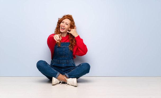 Redhead woman with overalls sitting on the floor making phone gesture and pointing front - Fotó, kép