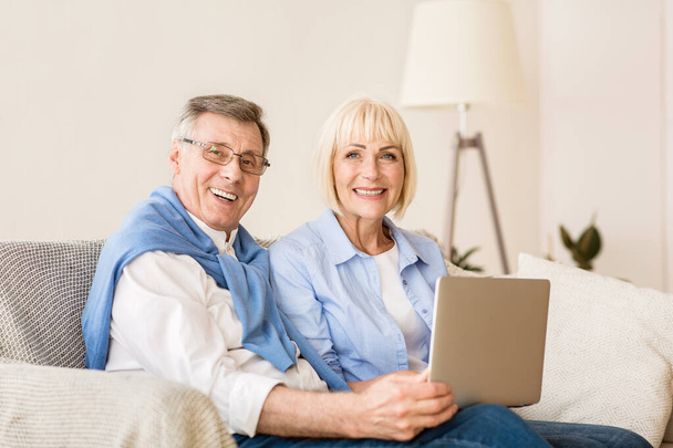 Joyful elderly couple with laptop smiling to camera - 写真・画像