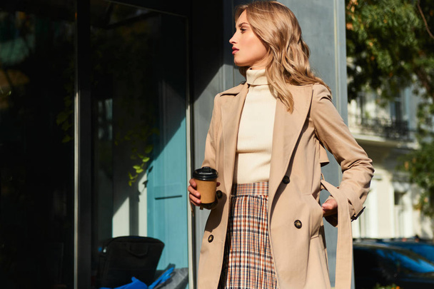 Vista lateral da menina loira elegante atraente em casaco bege com café para ir andando na rua da cidade
 - Foto, Imagem