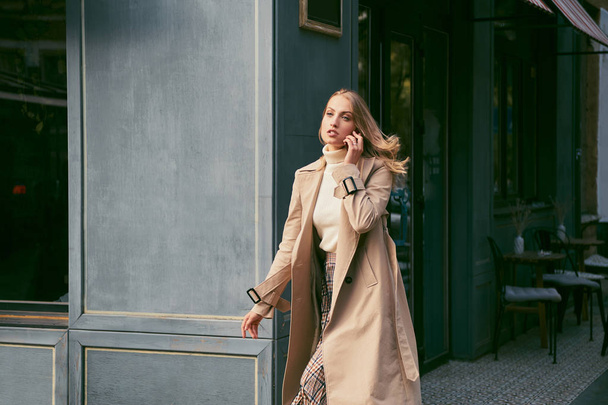 Beautiful blond girl in stylish trench coat talking on cellphone on street - Photo, Image