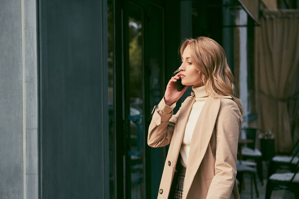 Vista lateral da menina loira atraente em casaco de trincheira falando pensativamente no celular ao ar livre
 - Foto, Imagem