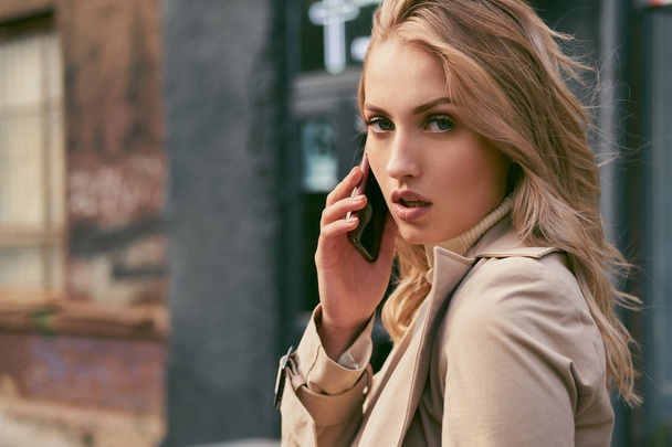 Attractive serious blond girl in trench coat talking on cellphone confidently looking in camera outdoor - Fotó, kép