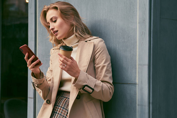 Atraktivní ležérní blond dívka v trenč kabát s kávou jít zasněně pomocí mobilního telefonu venkovní - Fotografie, Obrázek