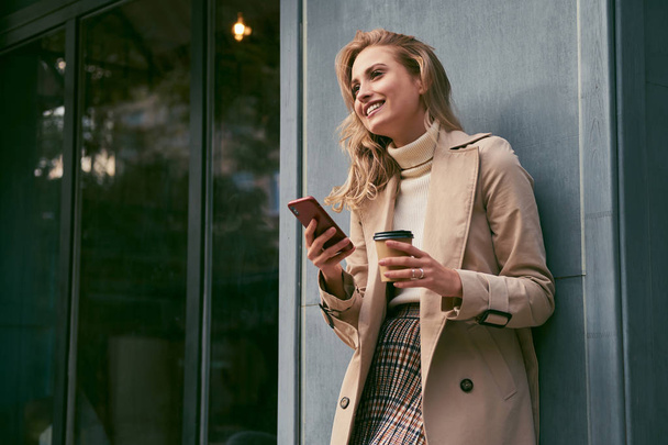 Attractive cheerful blond girl in trench coat with coffee to go happily using cellphone outdoor - Foto, Imagem