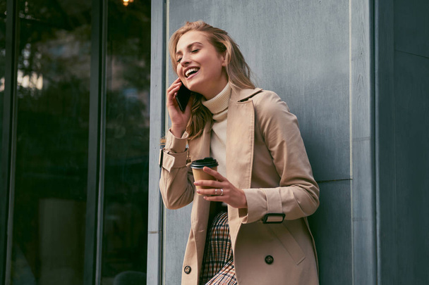 Hermosa chica rubia sonriente con elegante gabardina con café para ir felizmente hablando en el teléfono celular al aire libre
 - Foto, Imagen