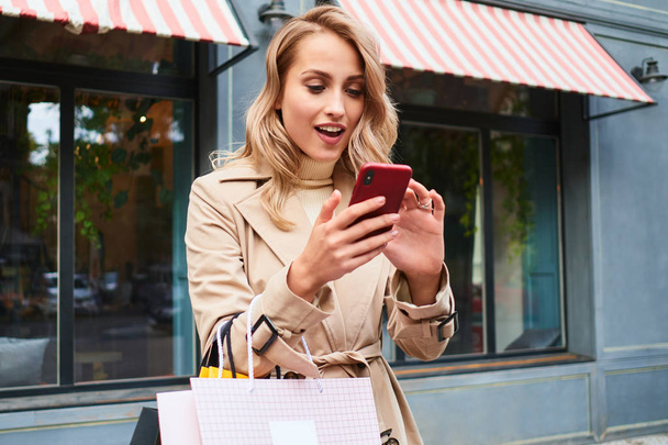 魅力的な驚きブロンド女の子でスタイリッシュなトレンチコートでショッピングバッグ喜んで使用携帯電話オンストリート - 写真・画像