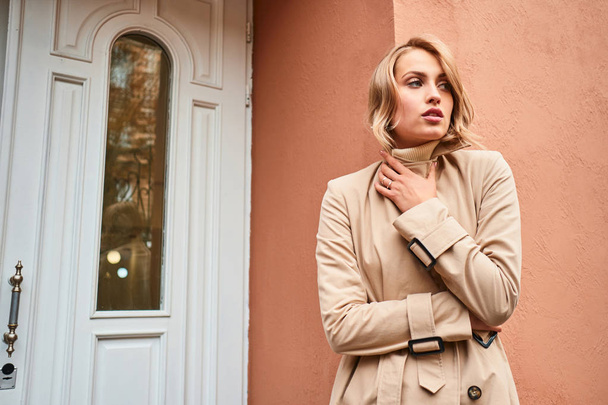 Aantrekkelijk casual blond meisje in beige trench jas bedachtzaam weg te kijken outdoor - Foto, afbeelding