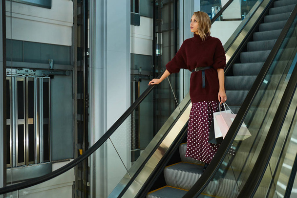 Bella ragazza bionda elegante con borse della spesa pensieroso guardando lontano sulla scala mobile nel moderno centro commerciale
 - Foto, immagini