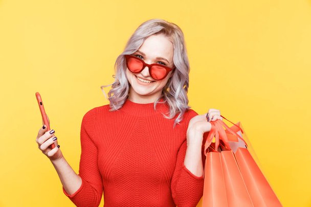 Blond woman in red glasses holding phone and bags isolated in the yellow studio. - 写真・画像