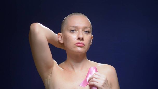 A bald woman who has survived cancer, holds in her hands a pink ribbon on a dark background. - Foto, Imagen