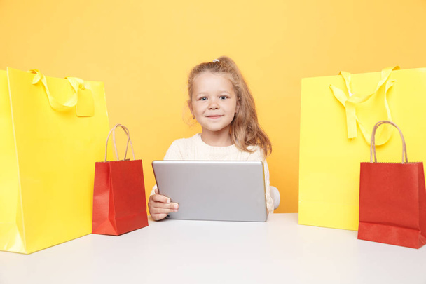 niña con la tableta y bolsas de compras sentado aislado sobre el fondo amarillo
. - Foto, imagen