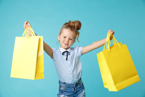 Little cute and funny girl holding big bright colored yellow paper bags in the blue studio. - Photo, Image