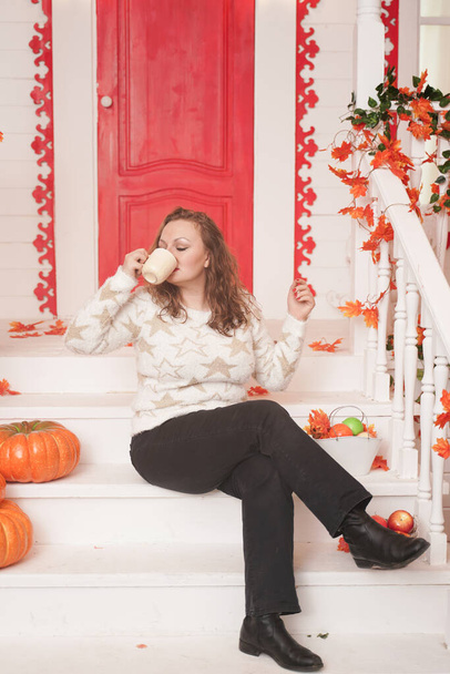 Αρκετά νέος συν μέγεθος γυναίκα σε ζεστό λευκό πουλόβερ πίνοντας τον πρώτο καφέ της ημέρας κάθεται στη βεράντα ενός όμορφου φθινοπωρινού σπιτιού με μια κόκκινη πόρτα - Φωτογραφία, εικόνα