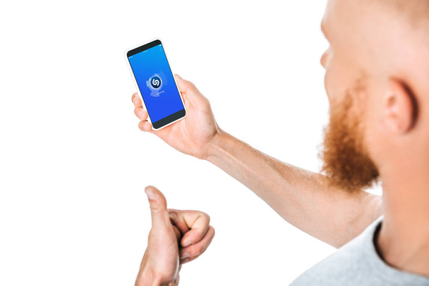 KYIV, UKRAINE - AUGUST 27, 2019: cropped view of man showing thumb up and looking at smartphone with shazam app, isolated on white - Фото, зображення