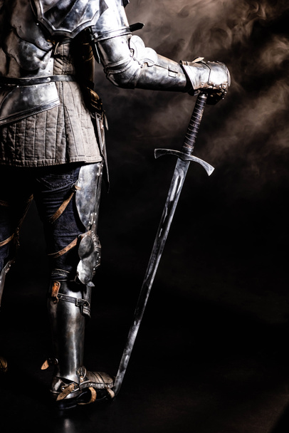 cropped view of knight in armor holding sword on black background  - Fotó, kép