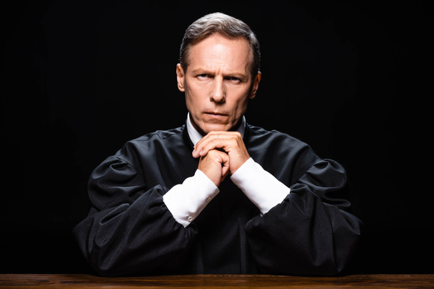 rechter in gerechtelijke robe zitten aan tafel en kijken naar camera geïsoleerd op zwart - Foto, afbeelding