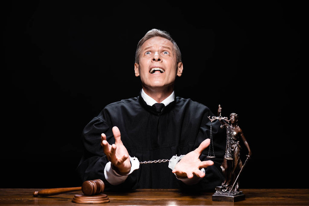 angry judge in judicial robe sitting at table with handcuffs and scream isolated on black - Photo, Image