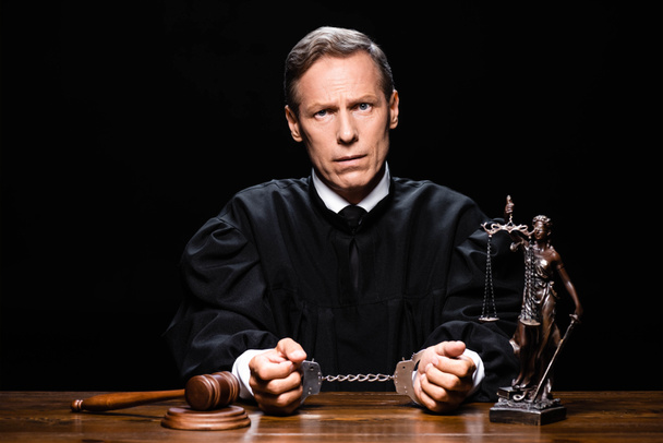 judge in judicial robe sitting at table with handcuffs isolated on black - Photo, Image